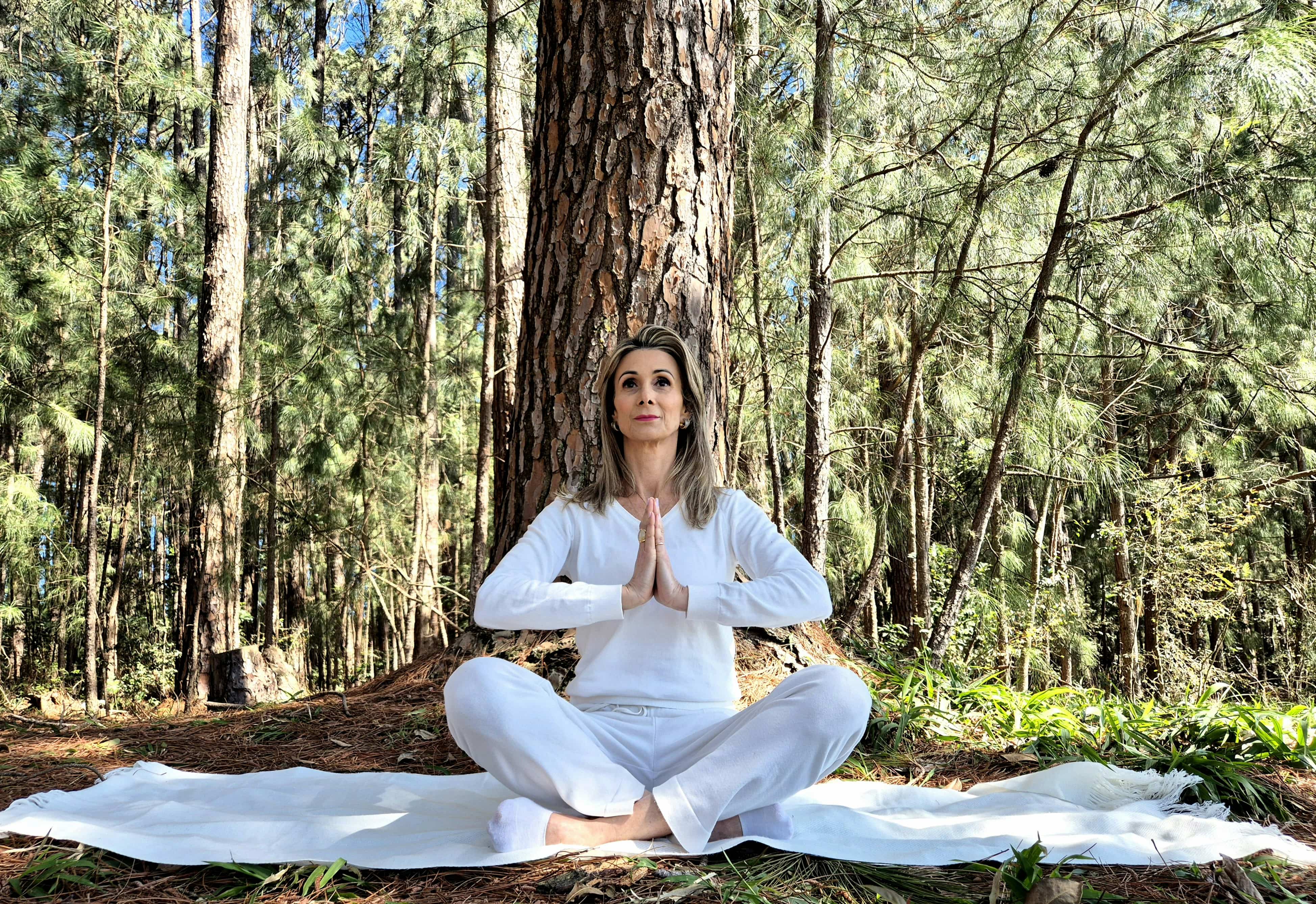 Fotografia de Juliane Fernandes fazendo com as mãos o sinal de Namaste próxima de árvores.